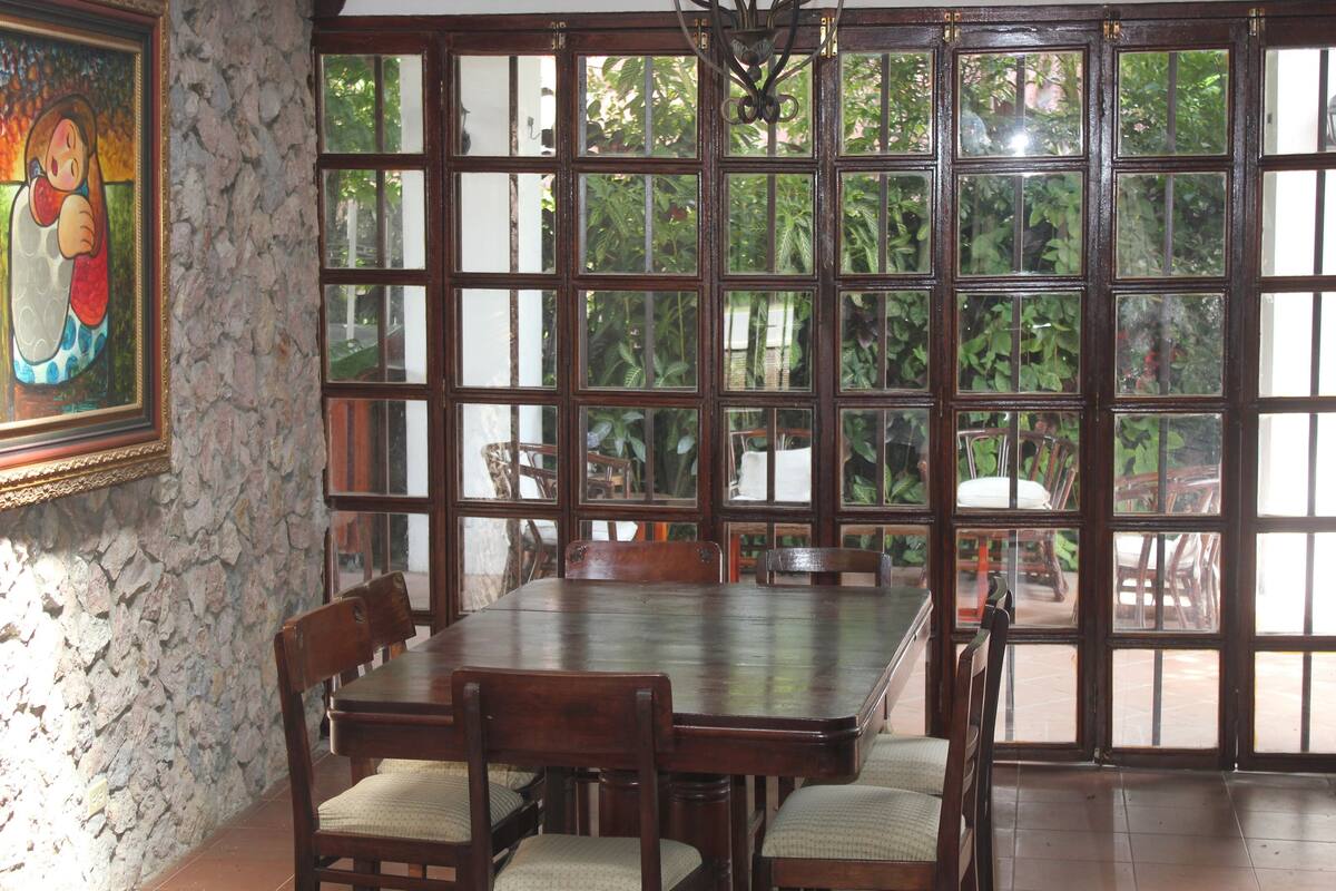 Comedor con gran mesa de madera y sillas, rodeado de ventanas amplias con vista a la vegetación.