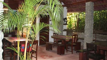 A dining area with wooden tables and benches, surrounded by stone pillars and a large palm tree.