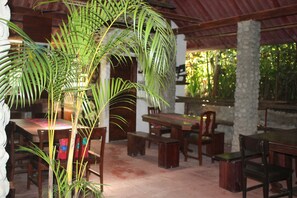 Un comedor con mesas y bancos de madera, rodeado de pilares de piedra y una palmera grande.