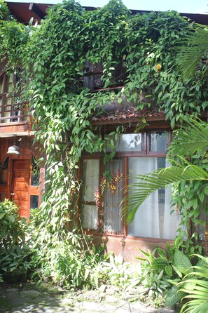 Una casa de madera con balcón, rodeada de abundante follaje y plantas.