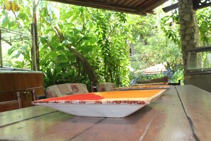 Una mesa de madera con superficie de baldosas, rodeada de sillas con cojines estampados, ubicada en un espacio exterior verde y frondoso con un pilar de piedra y una estructura de madera al fondo.
