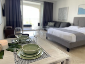 A neatly set dining table with plates, glasses, and a candle, a bed, and a view of the outdoors. - AquaBLU Ocean View Studio (Providenciales)