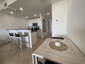 A modern kitchen with white cabinets, a microwave, and a coffee maker. There is a dining area with a table and chairs. - AquaBLU Ocean View Studio (Providenciales)