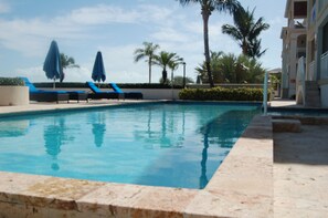 Une piscine entourée de palmiers et d’un bâtiment doté de balcons, avec des chaises longues et des parasols à proximité.