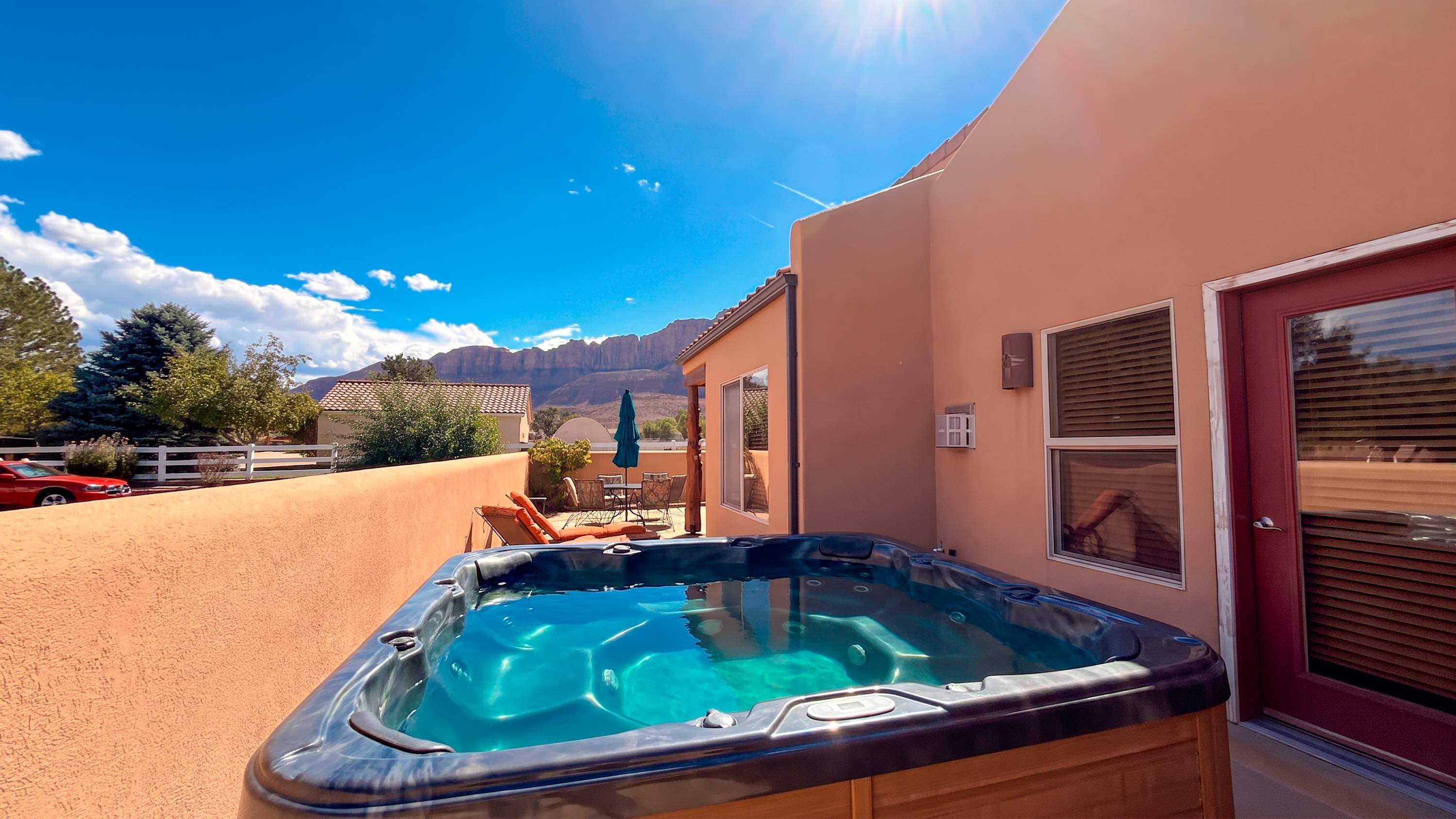 A hot tub in a backyard with a mountain view, a red car, and a white fence.