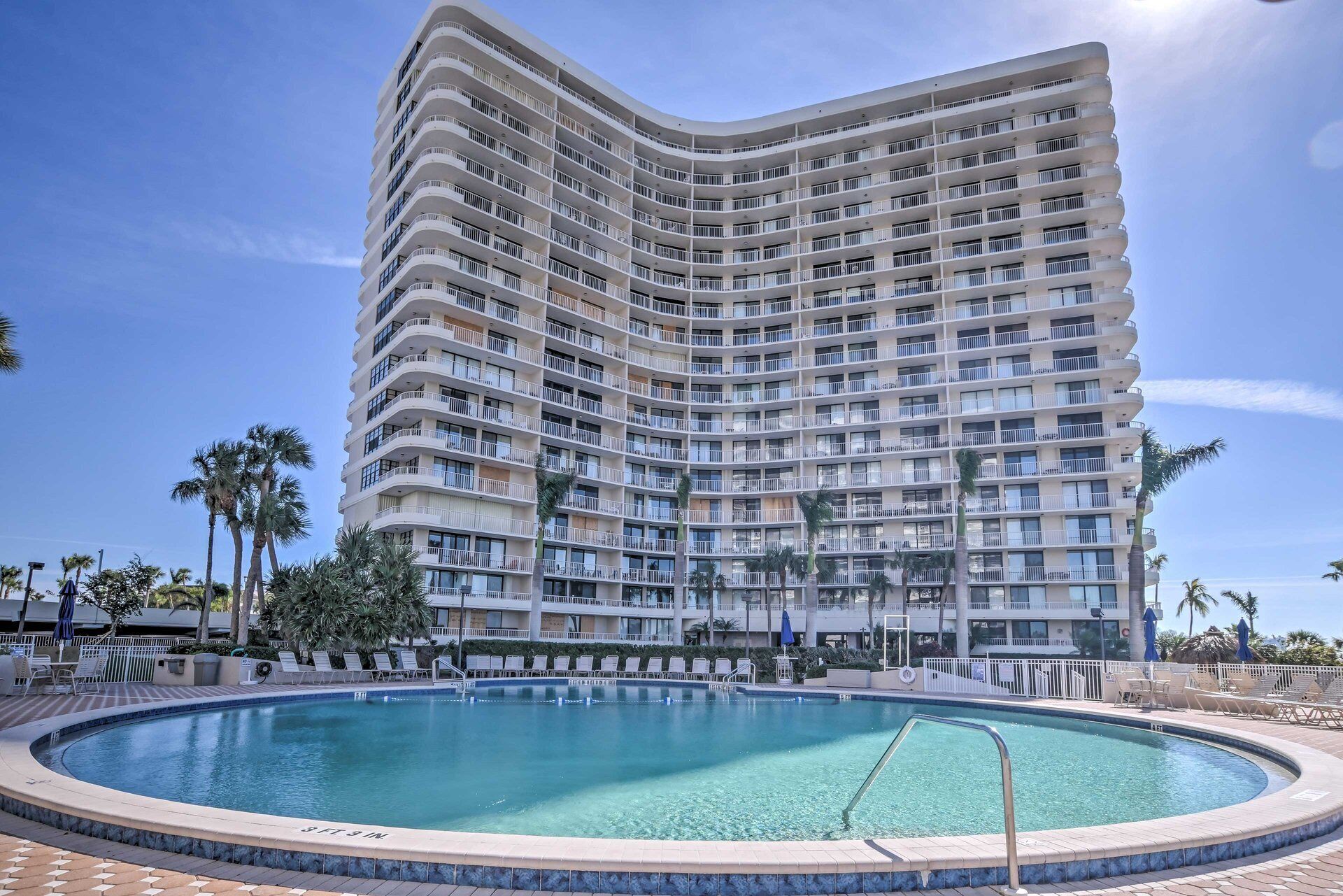 A multi-story hotel with a curved facade, a swimming pool, and palm trees.