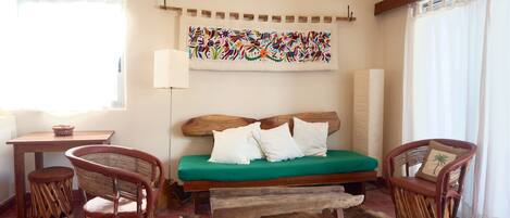 Quarto com cama de madeira, duas cadeiras de vime, uma mesinha e um tapete colorido na parede.
