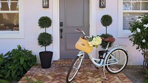 Ein weißes Fahrrad mit Korb und Blumen vor einem Haus mit grauer Tür und zwei Kübelbäumen.