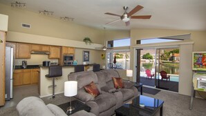 Sala de estar con sofá, mesa de centro y televisor. Zona de cocina con microondas, tostadora y armarios. Se observa un ventilador de techo y una vista a la piscina exterior.