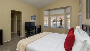 Un dormitorio con una cama grande, televisión y estantería. Hay ventanas con persianas y vistas a un puerto deportivo.