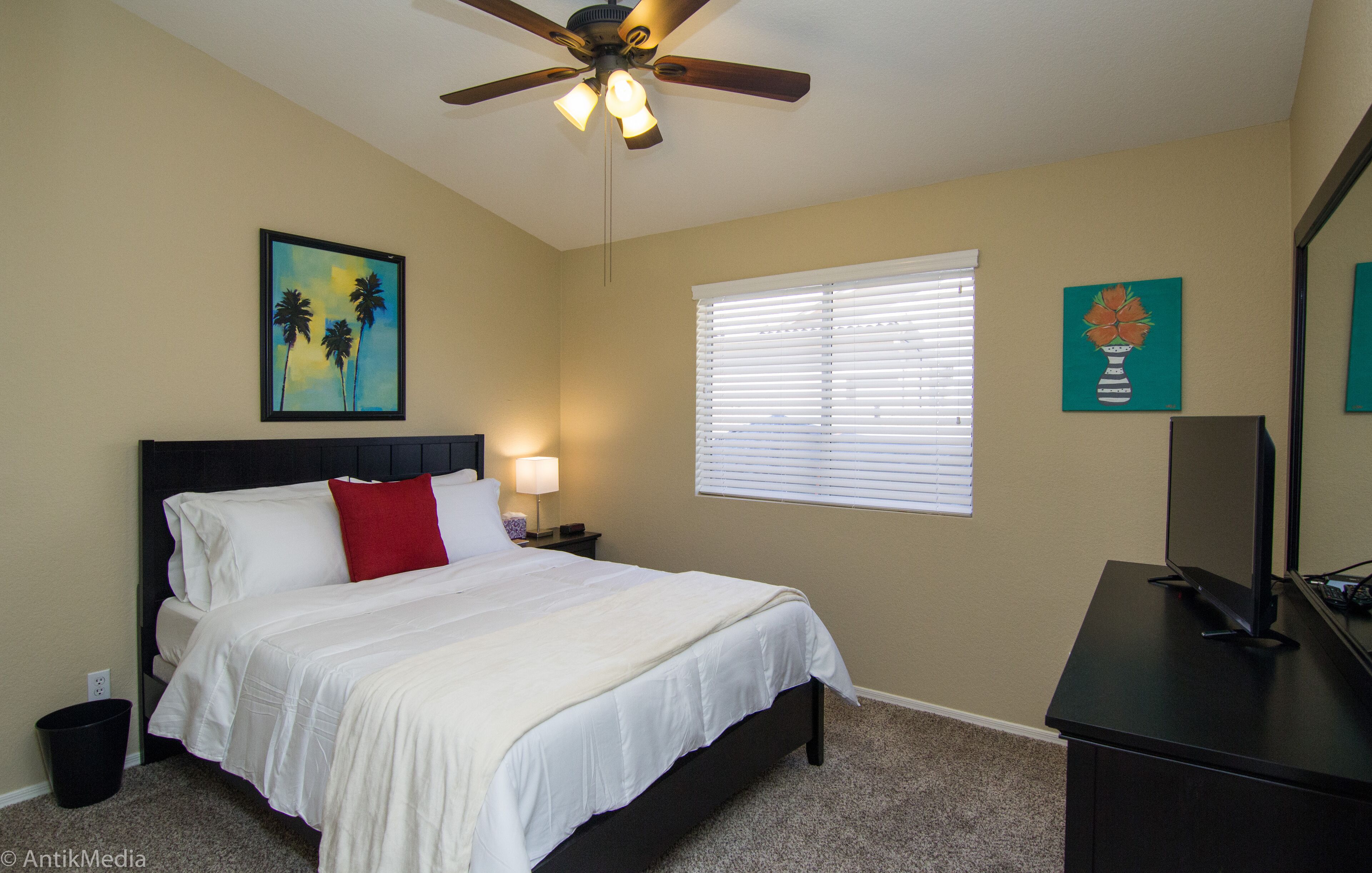 A bedroom with a bed, a ceiling fan, a window with blinds, and artwork on the walls.