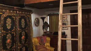A room with a wooden ceiling, a ladder, a patterned cabinet, a yellow armchair, and a wall clock.