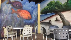 Ein Wandgemälde mit Fischen und einem Baum, Außenbereich mit Sitzgelegenheiten aus Metallstühlen und -tischen sowie ein Fliesenboden.