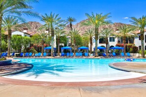 A swimming pool with blue lounge chairs and umbrellas, surrounded by palm trees and buildings. - Near Pool! Best Location! Single Story Poolside Villa across from Clubhouse, Pet Friendly! (C72) (La Quinta)