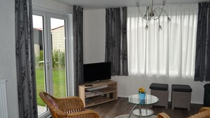 Una sala de estar con televisor sobre un soporte de madera, sillas de mimbre, una mesa de centro de vidrio y vistas a través de una puerta corrediza de cristal.