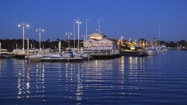 Uma marina com barcos atracados, um pequeno prédio com varanda e postes de iluminação à beira da água.