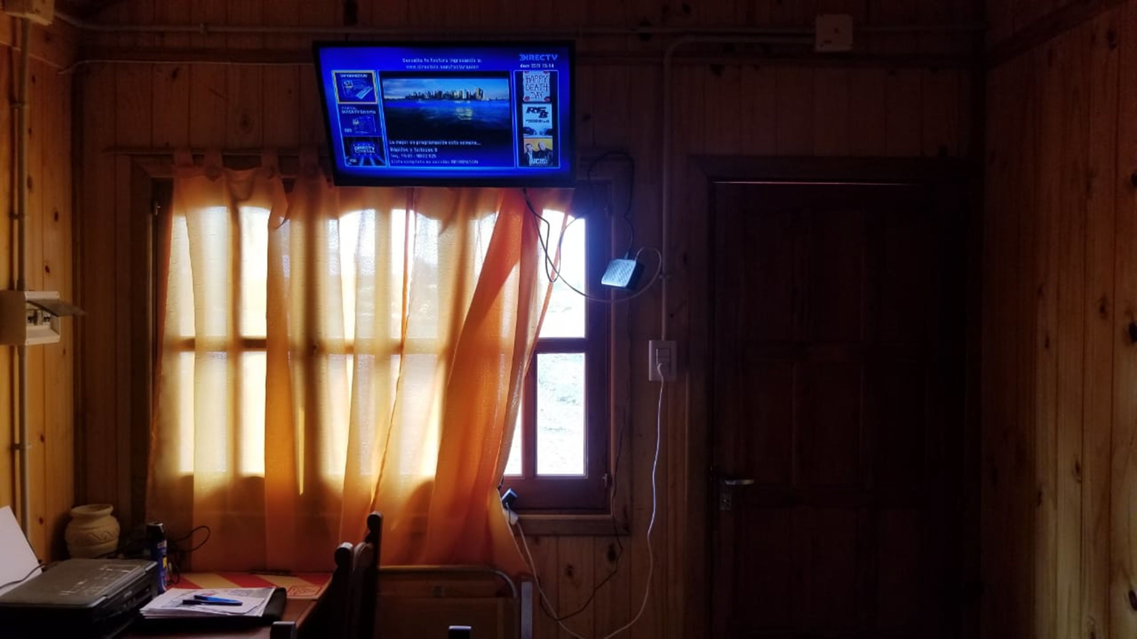 Une pièce avec un intérieur en bois, un téléviseur fixé au mur, un bureau avec un ordinateur portable et une fenêtre avec des rideaux.
