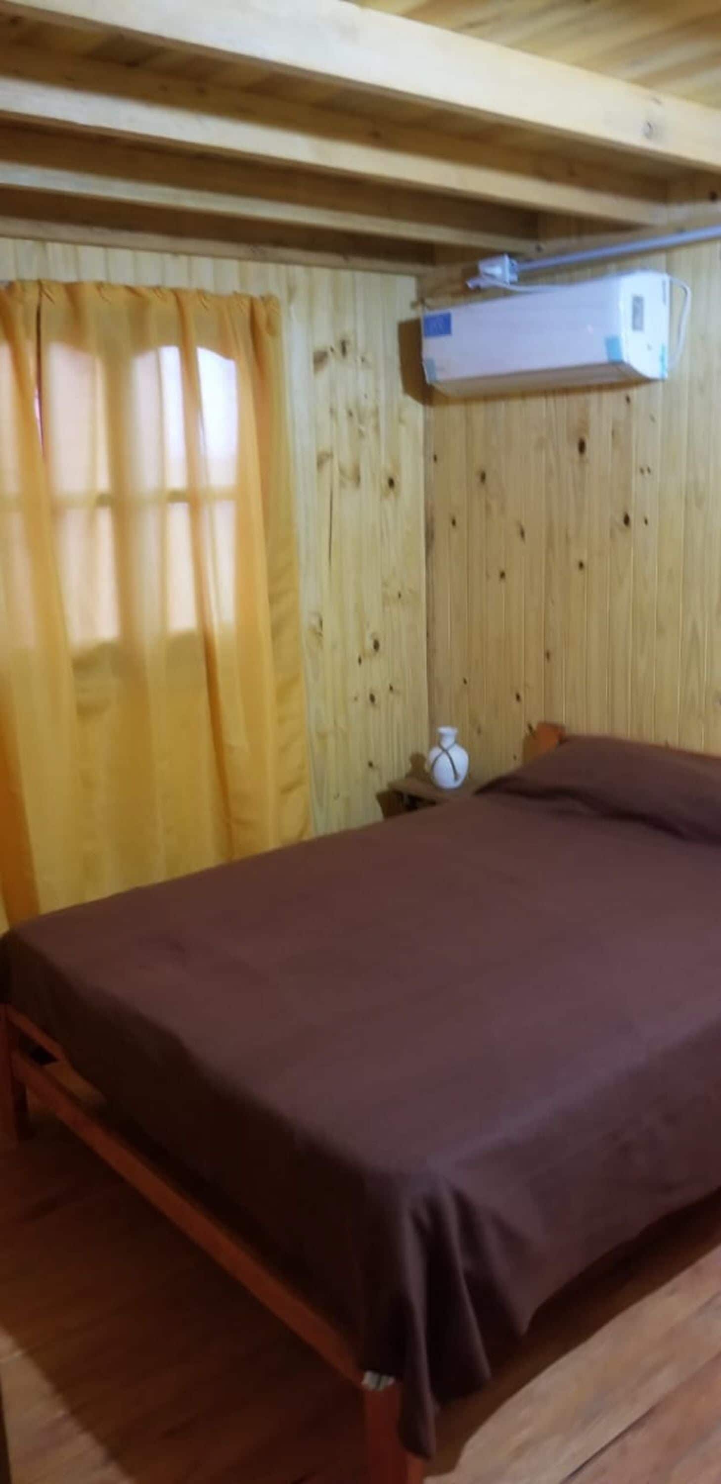 A wooden cabin room with a bed, curtains, a heater, and a small table with a cup.