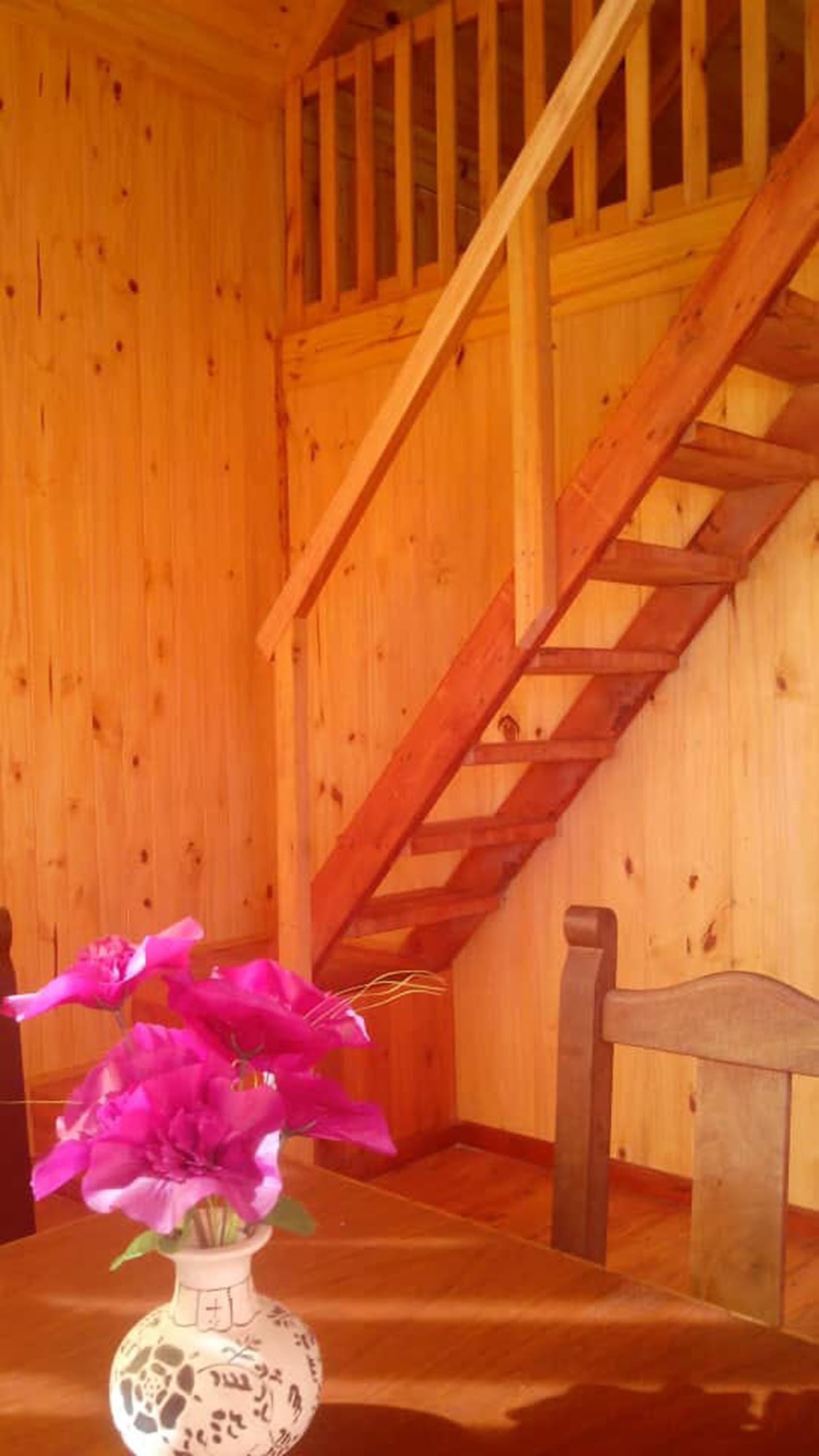 En interiör med träpanel, en trappa, en trästol och en vas med blommor på ett bord.