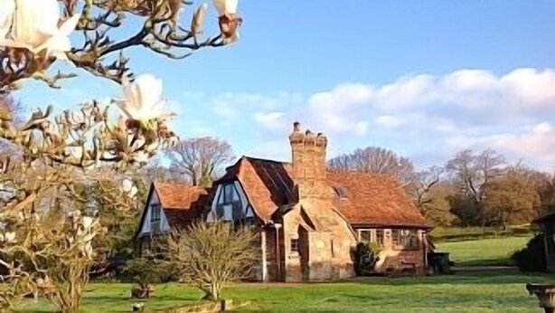A stone house with a sloped roof and a chimney, surrounded by a well-maintained lawn and flowering trees. - Outstanding Self Catering Cottage With Beautiful Gardens (Albourne)