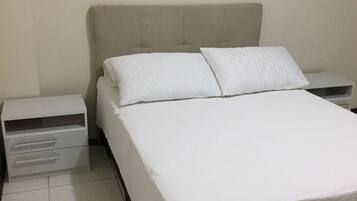 Uma cama com colcha branca e duas almofadas, uma mesa de cabeceira com gaveta e uma parede lisa.