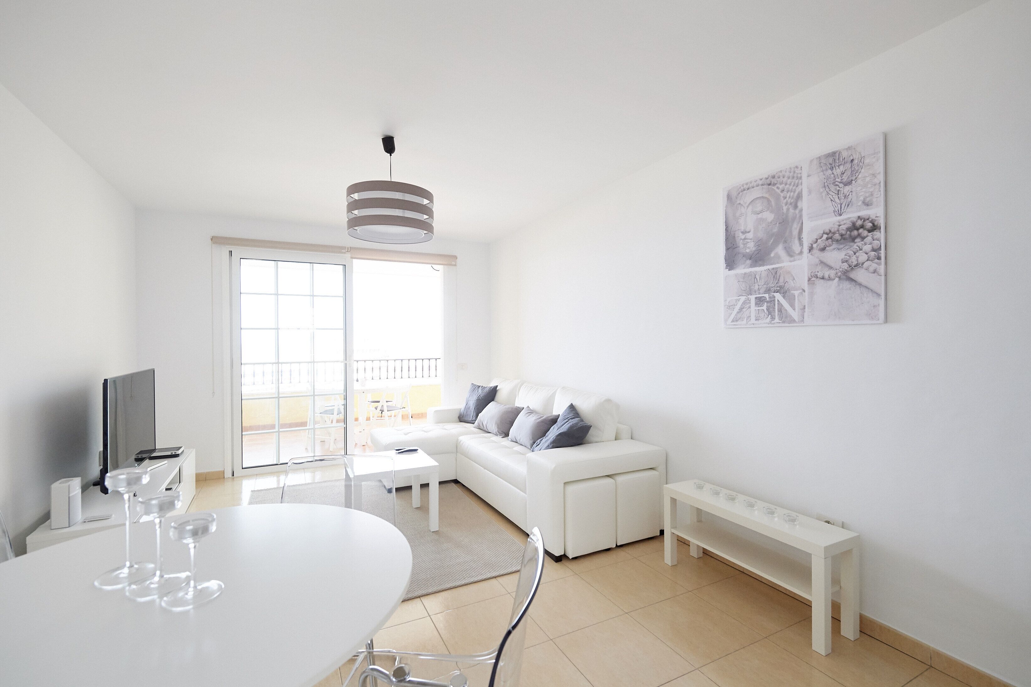 Ein modernes Wohnzimmer mit einem weißen Sofa, einem Esstisch mit Gläsern, einer Bank und einem großen Fenster.