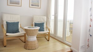 Une pièce comprenant deux fauteuils en osier avec des coussins, une petite table ronde et une grande fenêtre donnant sur la plage.
