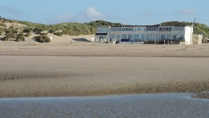 Un bâtiment situé sur la côte, doté d’un toit plat, de nombreuses fenêtres et bordé d’une plage de sable au premier plan.