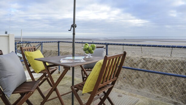 Une table surmontée d’un parasol blanc, deux chaises en bois avec des coussins, et un saladier de pommes vertes, le tout sur un balcon donnant sur une plage.