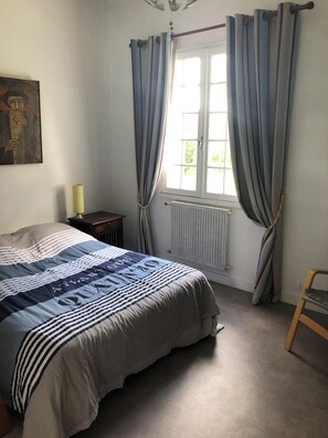 Una camera da letto con un letto, un termosifone, una finestra con tende, un tavolino con una lampada e una sedia.
