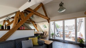 Un salon moderne avec un canapé gris, des poutres en bois, une table en bois et une grande porte-fenêtre coulissante donnant sur une terrasse extérieure.
