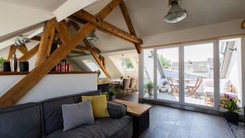 Ein modernes Wohnzimmer mit grauem Sofa, Holzbalken, einem Holztisch und einer großen Schiebeglas-Tür, die auf eine Außenpatio führt.