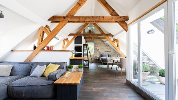 Un espace de vie moderne de style loft, avec des poutres en bois apparentes, un canapé d’angle gris, une table basse en bois et une grande porte-fenêtre coulissante donnant sur un espace extérieur.