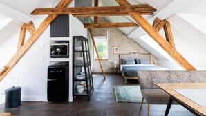 Une chambre à coucher aménagée dans un espace mansardé, avec des poutres apparentes, un lit, un canapé, une petite table et un coin cuisine équipé d’un réfrigérateur et d’étagères.