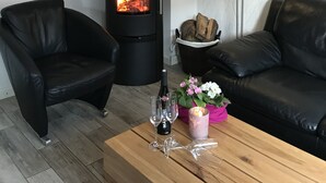 A modern living room with a wood-burning stove, black leather sofas, a wooden coffee table with wine glasses and flowers, and hanging light fixtures.