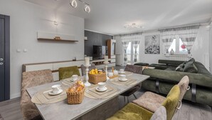 Ein modernes Wohnzimmer mit Essbereich, ausgestattet mit einer Sofagarnitur, einem Fernseher und einem Esstisch mit Unterlagen und einem Brotkorb.