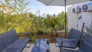 An outdoor seating area with two chairs and a table under a large umbrella, surrounded by greenery and a white wall with air conditioning units.