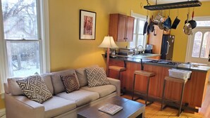 Ein Wohnzimmer mit einer Couch, einem Couchtisch und einer Küchenzeile mit Barhocker, Herd und hängenden Töpfen.