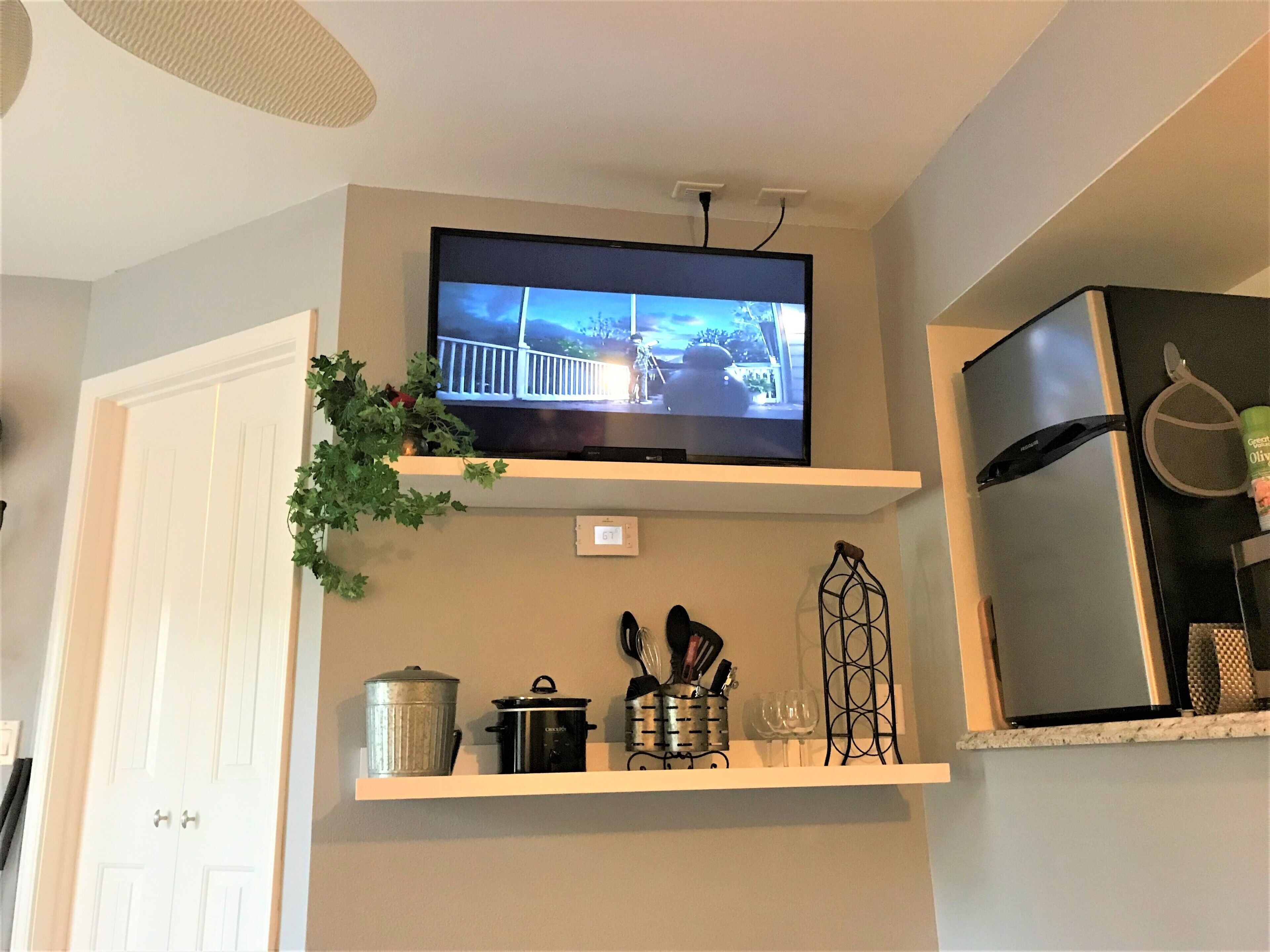 Una cocina con un televisor montado en un estante, un refrigerador y varios utensilios y utensilios de cocina sobre el mostrador.