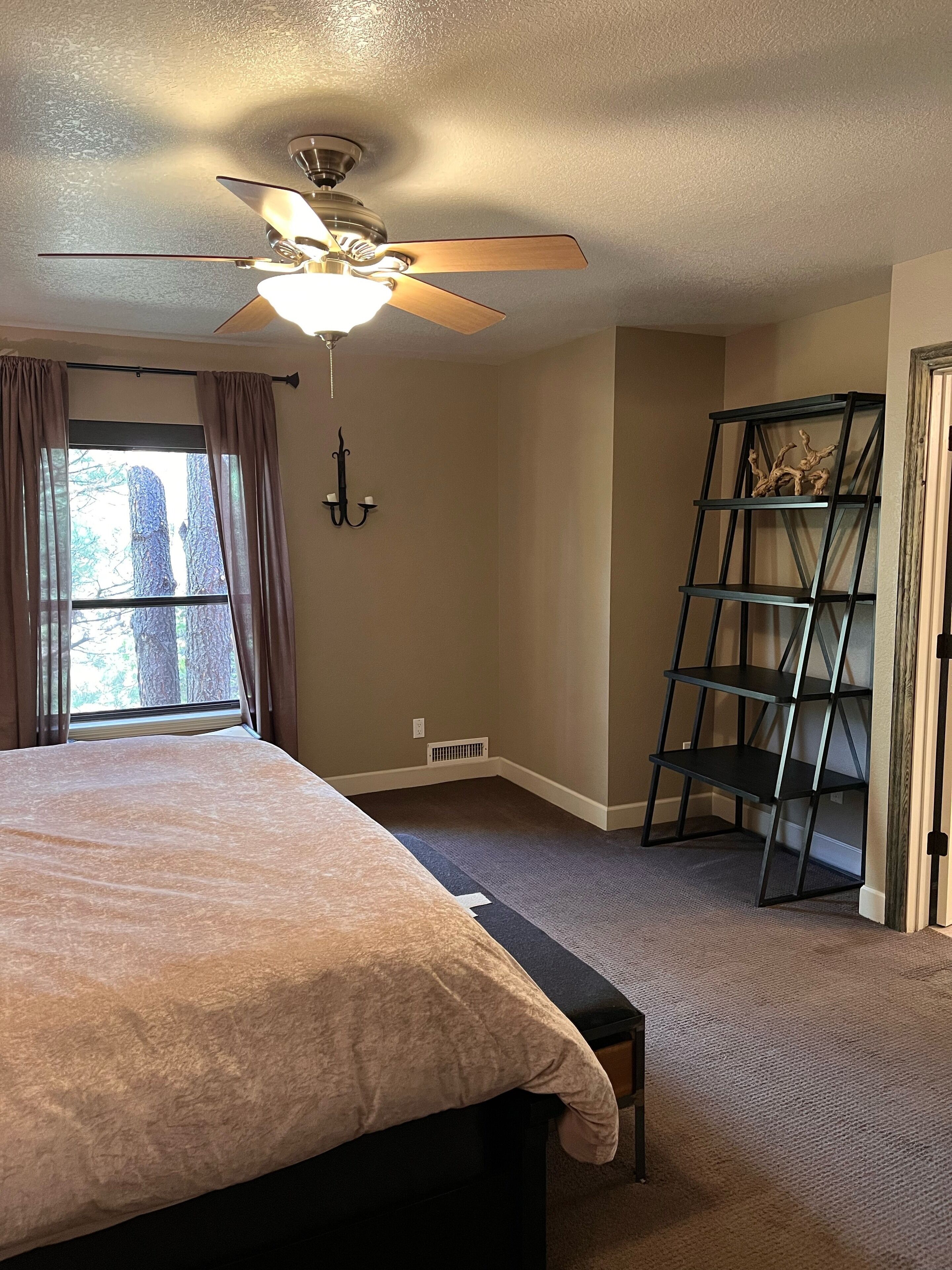 A bedroom with a bed, a ceiling fan, a window with curtains, a wall-mounted shelf, and a carpeted floor.
