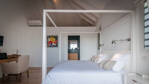 Una camera da letto con un letto grande, una scrivania con una sedia, un quadro appeso al muro e una zona cucina visibile attraverso un'apertura.