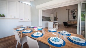 Zona pranzo con tavolo in legno apparecchiato per sei persone, pareti bianche e vista su cucina e soggiorno.