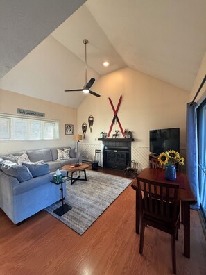 Ein Wohnzimmer mit Sofa, Couchtisch, Kamin, Fernseher und einem Essbereich mit Stühlen und einer Vase mit Sonnenblumen.