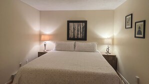 Une chambre avec un grand lit, deux tables de chevet avec des lampes, un banc au pied du lit et un tableau encadré au mur.