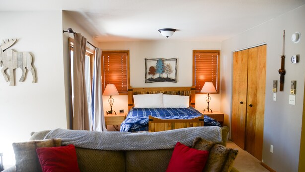 A bedroom with a bed, two bedside lamps, a wooden wardrobe, a wall-mounted artwork, and a sofa with red cushions. - Cozy large studio condo. (Winter Park)