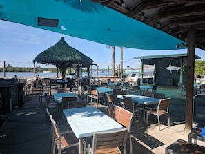 An outdoor dining area with tables and chairs, a thatched-roof structure, and a view of the water.