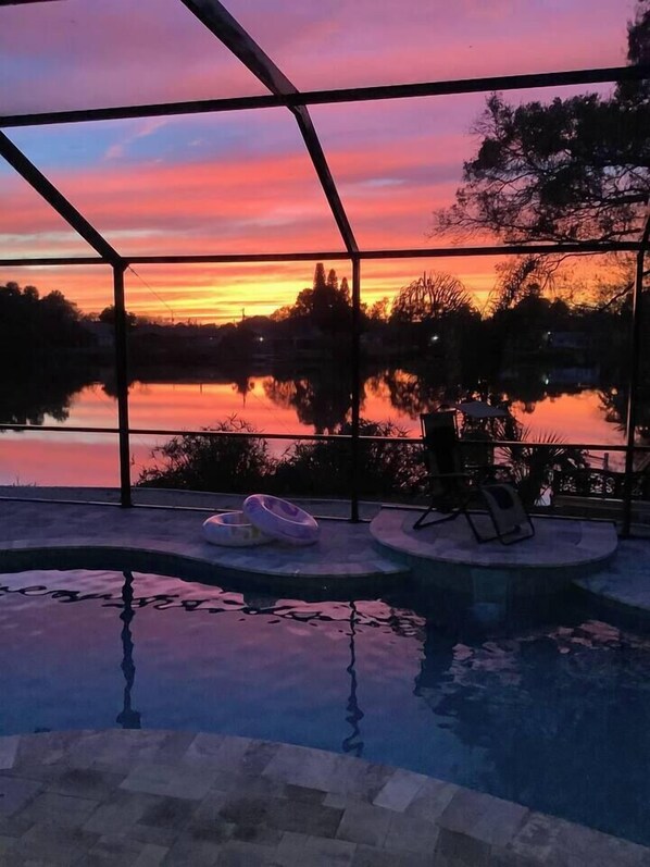 An indoor swimming pool with a view of a sunset over a body of water, surrounded by trees and a metal fence.