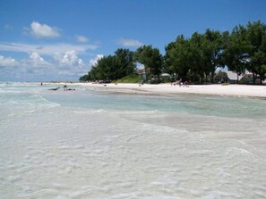 A beach with clear turquoise water, white sand, and a row of trees and buildings along the shore.