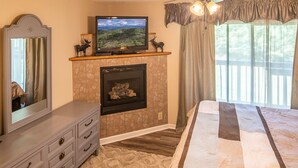 Un dormitorio con cama, cómoda, televisor, chimenea y ventanas amplias.