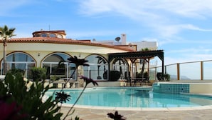 A swimming pool with a patio area, a covered seating area, and a building with arched windows and a tiled roof.